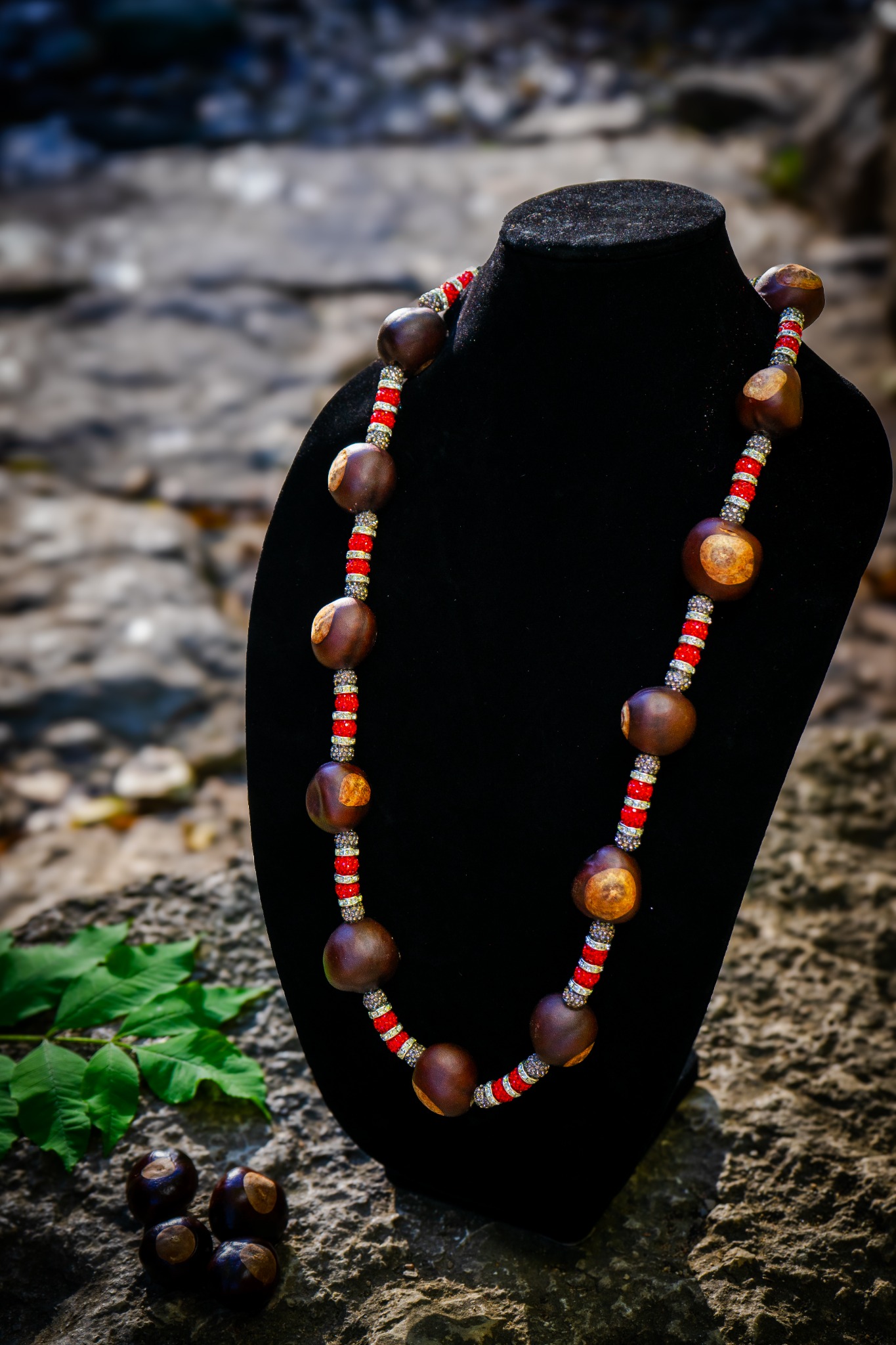 Handmade Buckeye necklace with red and gray rhinestone beads and silver-tone crystal spacers, displayed on a black velvet stand outdoors.