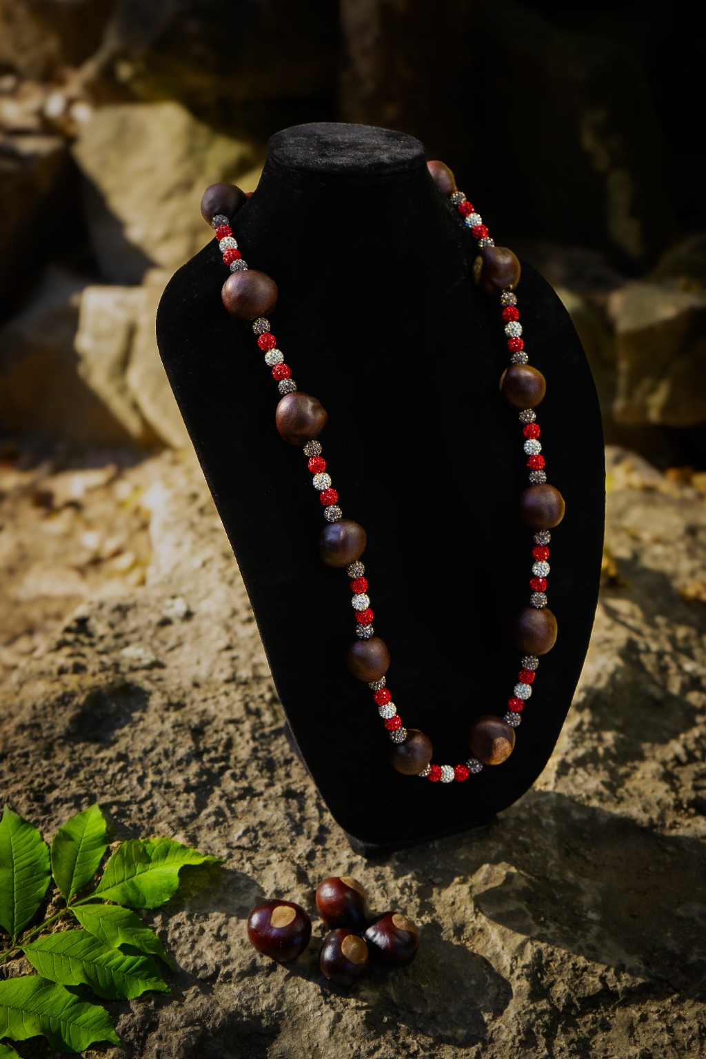 Handmade buckeye necklace with gray, red and white rhinestone beads, displayed on a black velvet bust outdoors.